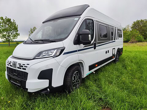 Weißer Fiat Ducato Camper auf grünem Feld mit Bäumen im Hintergrund.